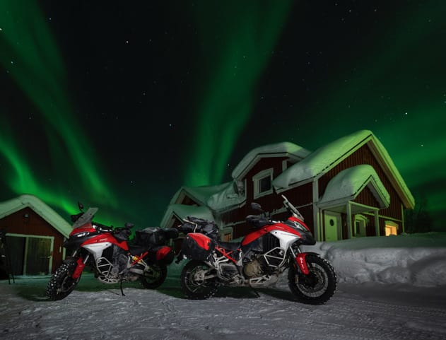 Zwei Motorräder vor verschneitem Haus und Polarlichtern