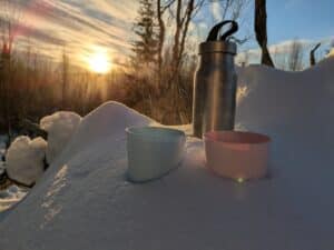 Thermoskanne und Tassen im Schnee bei Sonnenuntergang.